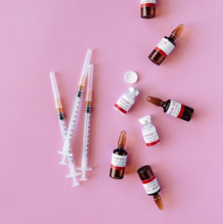 Foto von Spritzen und kleinen Flaschen, auf denen Covid-19-Vaccine steht auf pinkem Hintergrund